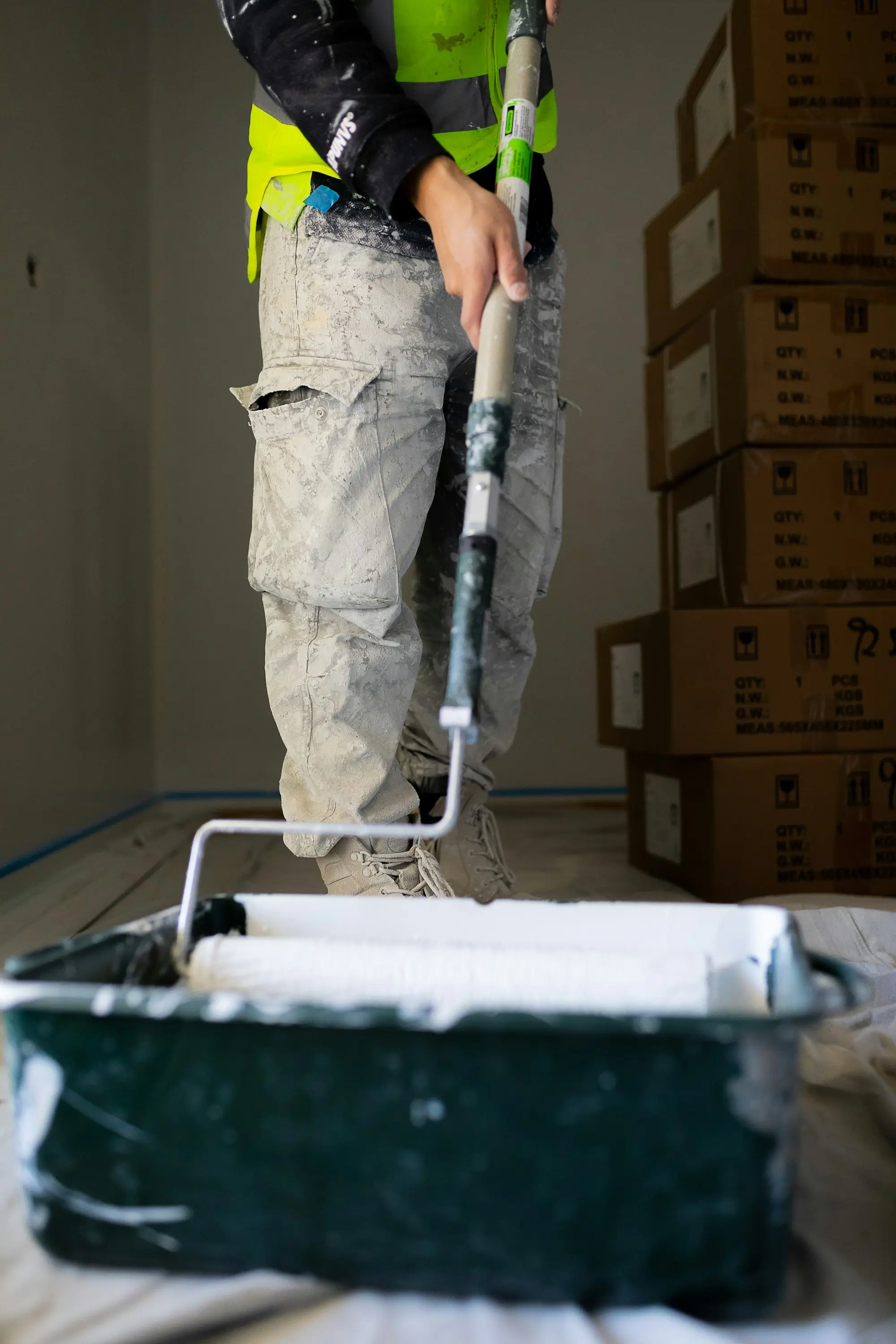 Painter in hi-vis rolling white paint onto a wall, paint tray in the foreground