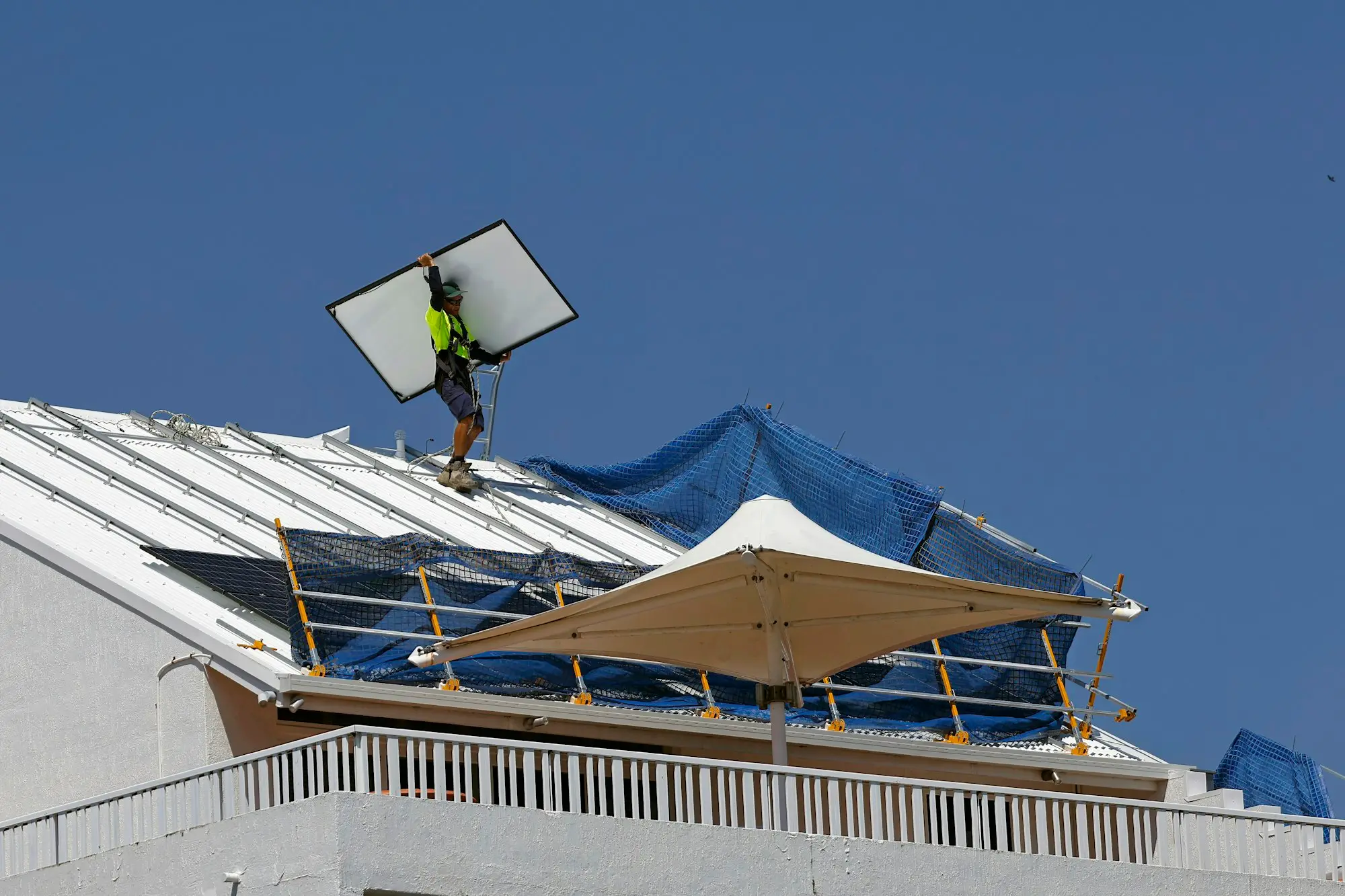 Tradie in hi-vis and harness carrying a roof panel across a metal roof in Cairns