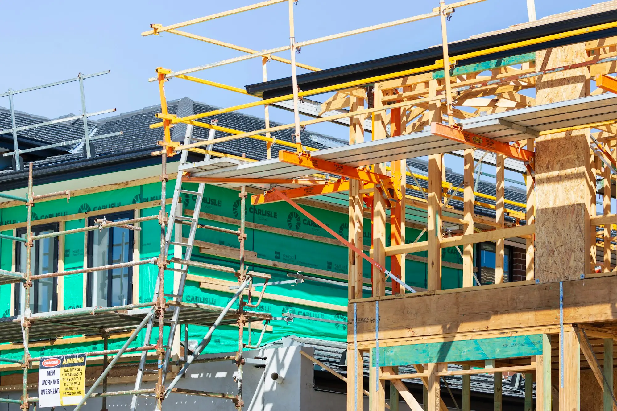Scaffolding and a ladder around a timber-framed new home with a men-working-overhead sign